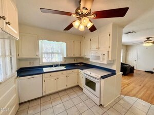 Kitchen with white cabinets, blue countertops, white appliances, tile flooring, and a ceiling fan with lights. There is a window above the sink and a view of a living area with wood flooring in the background.