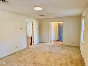 A spacious, empty room with beige carpet, white walls, two open doorways leading to other rooms, and windows letting in natural light. The ceiling has a round light fixture and there are minimal wall features.
