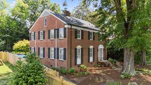 A two-story red brick house with white trim and black shutters, surrounded by trees and a wooden fence. The yard is landscaped with mulch and plants, and there is a small walkway to the side entrance.