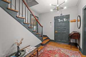 A hallway with a dark green front door, wooden staircase with painted risers, a vintage side table, a patterned rug, and a chandelier hanging from the ceiling. Light grey walls and wood floors are visible.