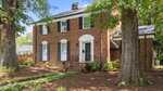 A two-story red brick house with black shutters, a white arched doorway, and a small front porch stands at 925 S Chestnut Street in Henderson. The yard features large trees, mulch, landscaped plants, and a wooden fence in the background.
