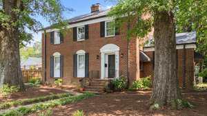 A two-story red brick house with black shutters, a white arched doorway, and a small front porch stands at 925 S Chestnut Street in Henderson. The yard features large trees, mulch, landscaped plants, and a wooden fence in the background.