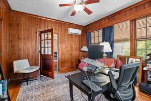 A home office with wood-paneled walls, a ceiling fan, a desk with computer equipment, an office chair, a white side chair, a red sofa, large windows with blue curtains, and a patterned rug on the floor.