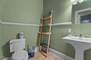 A small bathroom with green walls features a white toilet, a white pedestal sink, a ladder-style wooden shelf, a trash can, a toilet brush, and a rectangular mirror. The floor is covered with beige tiles.