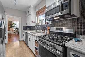 A modern kitchen with stainless steel appliances, gas stove, microwave, granite countertops, black tile backsplash, white cabinets, and a window above the sink. A dining area with plants is visible through an open doorway.