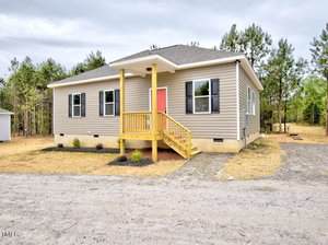 A single-story beige house at 48 S Grace Lane, Henderson, features a gabled roof, white trim, and a charming front porch with wooden steps. The red front door welcomes you, surrounded by trees and a gravel driveway.