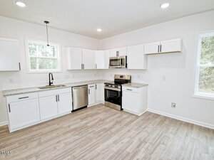 A modern kitchen with white cabinets, stainless steel appliances, a double sink under a window, light wood flooring, and recessed lighting. The space is clean and unfurnished with two large windows.