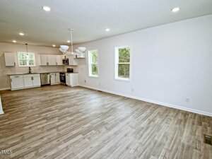 Open-concept room with light wood flooring, white kitchen cabinets, stainless steel appliances, and large windows letting in natural light. Ceiling lights and a modern chandelier hang from the white ceiling.