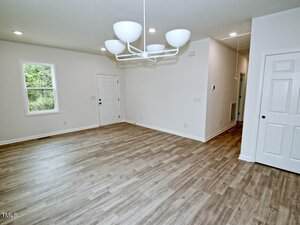 A spacious, empty room with light-colored wood flooring, white walls, a white door, a window, and a modern white chandelier hanging from the ceiling. There are two additional white doors visible.