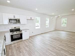 A spacious, unfurnished kitchen and dining area with light wood flooring, white cabinets, granite countertops, stainless steel appliances, and several large windows letting in natural light.