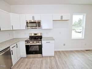 A modern kitchen with white cabinets, stainless steel appliances including a gas stove and microwave, granite countertops, light wood flooring, and a window showing a view outside.