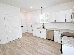 A modern kitchen with white cabinets, granite countertops, stainless steel appliances, a double sink under a window, light wood flooring, and two closed double-door pantries or closets.