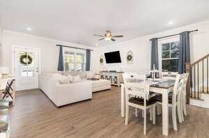 A modern open-concept living and dining area with light wood flooring, beige sectional sofa, wall-mounted TV, white dining table with six chairs, and large windows with gray curtains. A staircase is visible on the right.