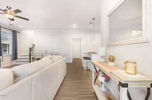 A modern living room with a beige sectional sofa, a ceiling fan, wood flooring, a console table with decor items, and a dining area with a white table and chairs. Stairs lead up to another level.