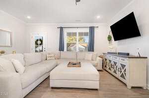 A bright living room with a large beige sectional sofa, matching ottoman, light wood floors, a wall-mounted TV, and a console with decorative items. A window with gray curtains lets in daylight.