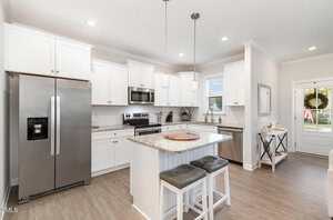 Bright, modern kitchen with white cabinets, stainless steel appliances, an island with two stools, and pendant lighting. There is a window above the sink and a door leading outside. The floor is light-colored wood.