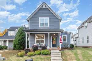 A two-story gray house with white trim at 203 Kipling Drive, Oxford, features a front porch with white railings, a wreath on the door, an American flag, and neatly trimmed shrubs. Steps lead from the sidewalk to the welcoming porch.