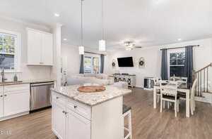 Open-concept kitchen and living area with white cabinets, granite island, stainless steel appliances, dining table, light wood floors, and a living room with a sectional sofa, TV, and large windows with gray curtains.