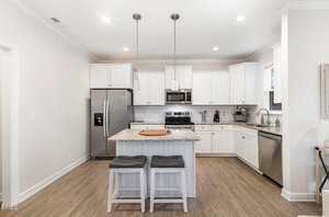 A modern kitchen with white cabinets, stainless steel appliances, a central island with a marble countertop, two gray cushioned stools, wooden flooring, and pendant lights hanging above the island.