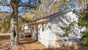 A small, single-story house with light-colored siding and a shingled roof sits among tall trees. A wooden porch is attached to the side, and the ground is covered with pine needles.