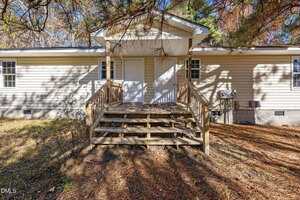 A wooden staircase leads to two white doors on the back of a beige, single-story house. The yard is covered with dry pine needles and surrounded by trees. An outdoor AC unit is mounted on the right side of the house.