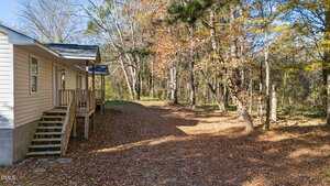 A beige house with a small porch and steps sits next to a dirt driveway, surrounded by trees with autumn leaves scattered on the ground.