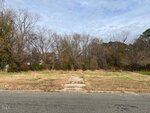 An empty lot at 501 Raleigh Road, Oxford, features overgrown grass and a narrow concrete path leading from the street to the center. Leafless trees and dense brush line the back edge of the lot under a cloudy sky.