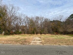 An empty lot at 501 Raleigh Road, Oxford, features overgrown grass and a narrow concrete path leading from the street to the center. Leafless trees and dense brush line the back edge of the lot under a cloudy sky.