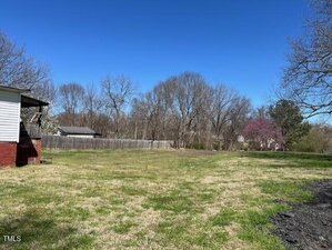 A grassy yard bordered by a wooden fence, with leafless trees in the background. Part of a white house with red brick foundation is visible on the left. The sky is clear and blue.