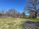 A grassy open lot at 0 Church Street, Oxford, with some bare patches and a section of dark soil, bordered by leafless trees and a wooden fence under a clear blue sky. A few background trees show early signs of spring blossoms.