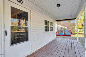 A white house porch with a wooden floor, white siding, a screen door, a double window, a ceiling light, and a colorful cushioned porch swing. Trees and a yard are visible in the background.