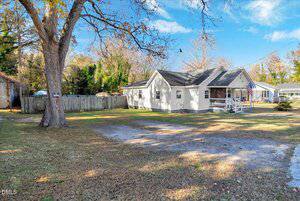 A single-story gray house with a front porch and American flag, set on a grassy lot with scattered trees and a gravel driveway. A wooden fence runs along the left side and other houses are visible in the background.