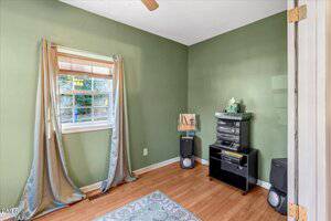 A small room with green walls, wood flooring, a window with draped curtains, a stereo system on a black stand, two speakers, and a tissue box on top. Part of a door and a rug are visible in the foreground.