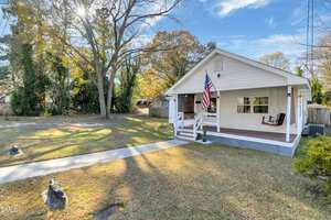 A small white house with a covered front porch, American flag, porch swing, and steps leading to a sidewalk. The yard is grassy with a few trees and some fallen leaves under a blue sky.