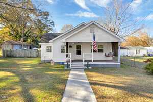Charming single-story white house at 509 N Clark Street, Henderson, featuring a covered front porch with an American flag and swing chair. Concrete walkway leads to the entrance, with a grassy yard, trees, and outbuildings in the background.