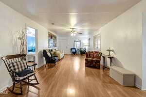 A spacious living room with light wood floors, white walls, several chairs, a small sofa, a ceiling fan with lights, and a side table. A bedroom is visible through an open doorway on the left.