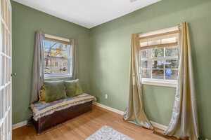 A small room with green walls, wood flooring, two windows with curtains, and a bench with cushions beneath one window. Daylight enters through the windows, and a rug is partially visible on the floor.