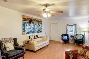 A living room with wood flooring, a ceiling fan with lights, and a large abstract painting on the wall. The seating includes a beige loveseat, armchairs, a side table, and a floor lamp near a curtained window.