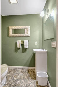 A small bathroom with green walls features a white vanity and sink, a wall-mounted mirror, a shell-framed decorative mirror, two towels on a rack, a white trash bin, and pebble-patterned floor tiles.