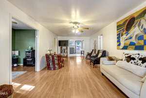 A spacious living room with light wood flooring, white walls, a white couch, patterned armchairs, ceiling fans, a large painting, and view into a green-walled room and kitchen in the background.