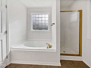 A white bathroom featuring a built-in bathtub with a gold faucet, a frosted glass window above, and a separate shower with a glass door and gold trim. The floor is covered with brown tiles.