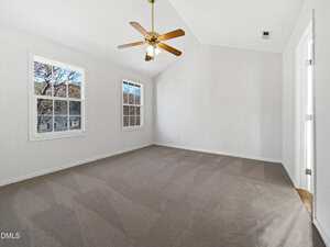 A spacious empty room with beige carpet, white walls, two large windows, a ceiling fan with lights, and two doors; sunlight streams through the windows, highlighting the clean, neutral interior.