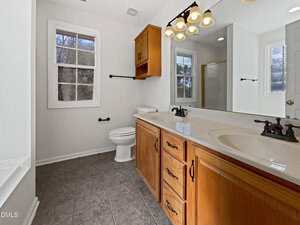 A bathroom with a double sink vanity, wooden cabinets, a toilet, a window, and a mirror above the sinks. The room has gray tile flooring and overhead lighting with four bulbs.