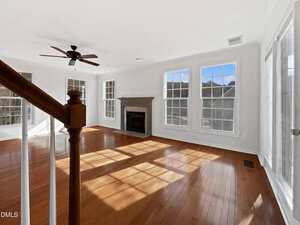 A bright, empty living room with large windows, hardwood floors, a ceiling fan, and a fireplace. Sunlight streams through the windows, casting shadows on the floor. A wooden staircase banister is visible in the foreground.