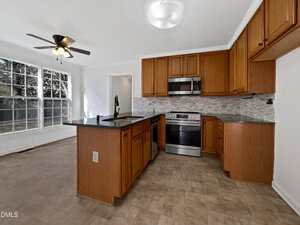 Kitchen with wood cabinets, stainless steel appliances, and a central island with a black countertop; tile flooring, backsplash with a gray pattern, and large windows letting in natural light. Ceiling fan and light fixture visible.