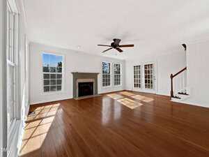 A spacious, empty living room with hardwood floors, a fireplace, large windows letting in natural light, a ceiling fan, and a staircase in the corner. French doors are visible on one wall.