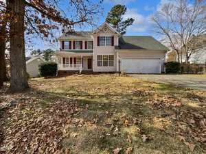 Two-story suburban house with white siding, red shutters, and a large front yard covered with fallen leaves. There is a three-car garage on the right and several trees with sparse leaves in the yard and surrounding area.