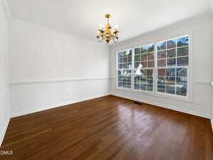 An empty room with white walls, a hardwood floor, a brass chandelier, and a large window showing a suburban street and trees outside. Natural light fills the space.