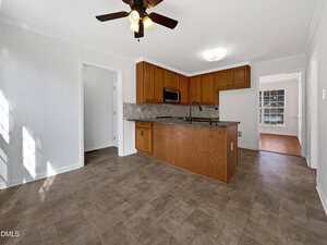 A kitchen with brown cabinets, black countertops, a built-in microwave, and tile flooring. There is a ceiling fan above, an open doorway to the left, and another room visible through a doorway on the right.