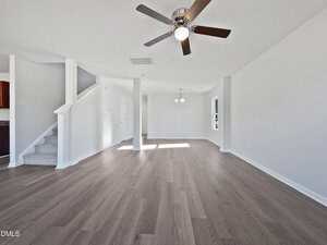 Spacious living area with light gray walls, wood-look flooring, ceiling fan with lights, and white staircase. Two pendant lights hang near a window, and natural light enters from multiple angles.